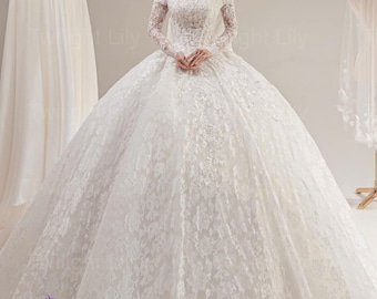 Puede incluir: Un vestido de novia blanco con hombros descubiertos, mangas largas de encaje y una falda amplia. El vestido presenta intrincados detalles de encaje y se exhibe en un estudio con otros vestidos en perchas.