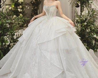 Puede incluir: Un vestido de novia blanco con un corpiño sin tirantes y una falda completa. La falda está hecha de capas de tul y está decorada con lentejuelas. El vestido es perfecto para una novia que quiere un look clásico y elegante.