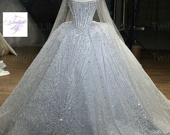 Puede incluir: Un vestido de gala blanco con corpiño estilo corsé, escote cuadrado y falda amplia, todo decorado con lentejuelas y cristales plateados en un patrón de estallido estelar. Un velo transparente a juego cae sobre los hombros.