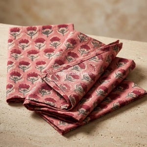 Op de afbeelding: Opgevouwen roze bloemen servetten met een herhalend patroon van rode bloemen en grijze bladeren. De servetten zijn gemaakt van een zachte stof en liggen op een lichtgekleurd oppervlak. De tekst "Jaipur Table Linen" is zichtbaar.