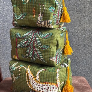 Peut inclure: Une pile de trois trousses rectangulaires en tissu matelassé vert olive, ornées d'imprimés de zèbres, de léopards et de feuillages tropicaux. Chaque trousse est dotée d'un pompon jaune vif attaché à la tirette de la fermeture éclair.