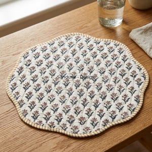 May include: A round, quilted fabric placemat with a scalloped edge featuring a repeating floral pattern of small pink flowers and dark green leaves on an off-white background. The border is a yellow and white striped fabric. Text reads Sandune Design.
