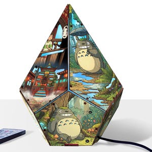 May include: A multi-faceted, diamond-shaped lamp featuring vibrant, detailed illustrations from Studio Ghibli films. The panels display scenes with Totoro, No-Face, and traditional Japanese architecture. A black power cord and remote are visible.