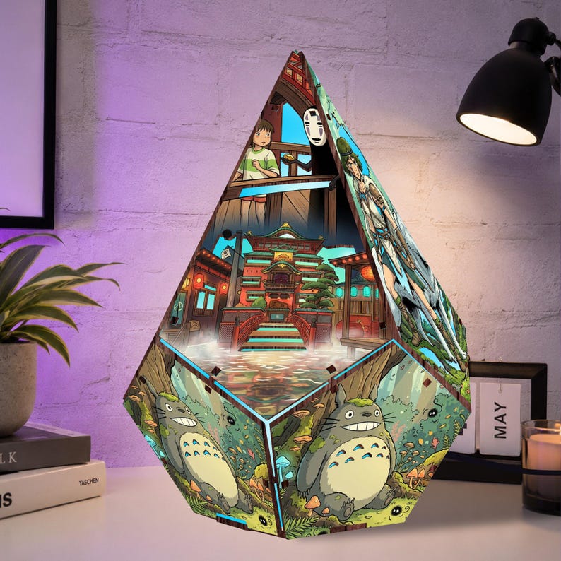 May include: A pyramid-shaped lamp featuring colorful illustrations from animated films. The design includes a bathhouse scene with a girl and a spirit, a character with a white mask, and a large grey creature with a white belly sitting in a forest.