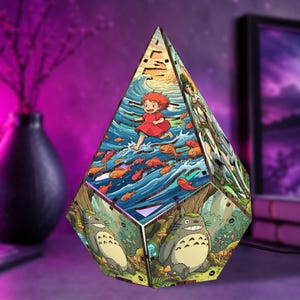 May include: A pyramid-shaped wooden lamp featuring colorful illustrations of animated characters. One side depicts a girl running through ocean waves with orange fish, while another side shows a large grey creature in a forest setting.