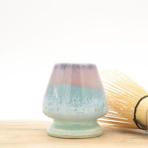 Könnte beinhalten: Eine Keramik-Matcha-Schale mit einem mehrfarbigen Farbschema aus Lila, Rosa und Blaugrün. Neben der Schale liegt ein Bambusbesen. Die Schale hat eine abgerundete Form mit breitem Boden und ausgestelltem Rand und ist etwa 10 cm hoch.