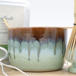 May include: A matcha tea set featuring a ceramic bowl with brown and green glaze, a bamboo whisk, a metal sieve, and a package of tea. The bowl has a unique drip glaze design. The tea package has the text "Simon Levelt".