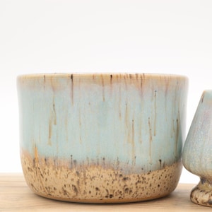 May include: Two ceramic plant pots with a turquoise glaze and a speckled brown base. The larger pot is cylindrical, while the smaller one has a unique, flared shape. Both have a dripped glaze effect, adding to their handmade aesthetic.