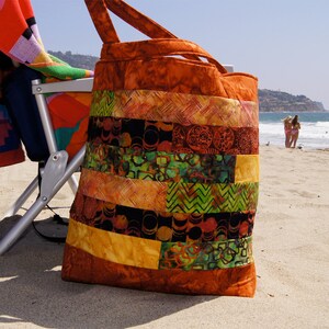 May include: A large, patchwork tote bag with a brown, orange, yellow, green, and black color scheme. The bag is made of fabric and has a wide, flat bottom.