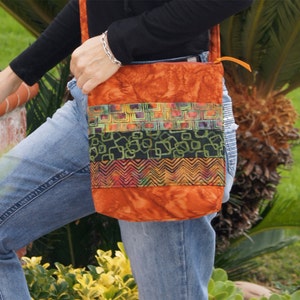 Può includere: Una borsa a tracolla in tessuto marrone con un motivo geometrico multicolore. La borsa ha una tracolla lunga e una chiusura a lembo.