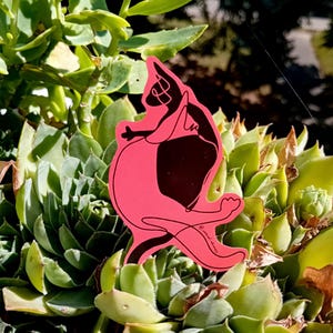 May include: A pink sticker featuring a black and white cat illustration. The cat is in a playful pose, with its body outlined in black and a solid black patch. The sticker is set against a backdrop of green succulent plants.