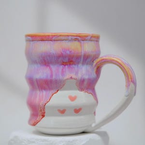 Puede incluir: Una taza de cerámica hecha a mano con un diseño ondulado y texturizado. La taza presenta una vibrante combinación de colores rosa, morado y amarillo, con una base blanca adornada con tres pequeños corazones rosas. El asa es blanca y amarilla.