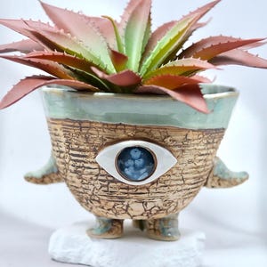 Puede incluir: Una maceta de cerámica con un acabado beige texturizado y agrietado, con un motivo de ojo azul central. El recipiente presenta pequeños brazos y pies que sobresalen, con un borde verde claro liso. Una suculenta rojiza y verde está dentro.
