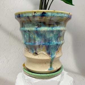May include: A ceramic plant pot with a unique, textured design. The pot features a cream-colored base, transitioning to a turquoise and blue glazed upper section. A matching green saucer sits beneath the pot, and a plant with green leaves is inside.