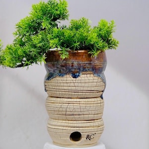 May include: A decorative ceramic planter with a layered design, featuring a brown and blue glazed top and a textured, off-white base. The planter holds a vibrant green, artificial plant. The base has the letters "RC" on it.