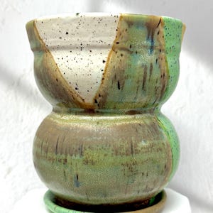 Op de afbeelding: Een handgemaakte keramische plantenbak met een uniek tweekleurig ontwerp. De plantenbak heeft een gespikkelde witte, bruine en groene glazuur. De bijpassende schotel is inbegrepen. Deze decoratieve pot is ideaal voor kleine planten.