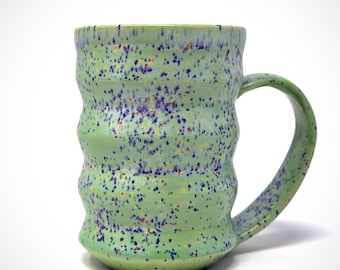Puede incluir: Taza de cerámica hecha a mano con un esmalte verde claro y detalles moteados en morado, amarillo y azul oscuro. La taza tiene un cuerpo acanalado y texturizado y un asa curva.