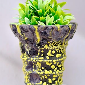 Könnte beinhalten: Ein dekorativer Keramik-Pflanztopf mit einer strukturierten schwarzen und gelben Außenseite, gefüllt mit leuchtend grüner Kunstpflanze. Der Topf hat ein einzigartiges, organisches Design, ideal für einen Hauch von Natur.
