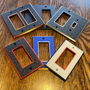 May include: A collection of decorative light switch covers in various colors. The covers feature a textured black surface with colored frames in red, blue, gold, and silver. The covers are designed for single and double switch configurations.