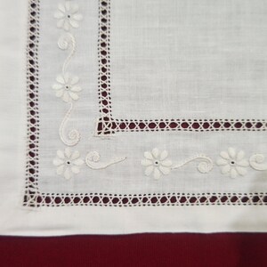 Op de afbeelding: Wit linnen textiel met geborduurde bloemendetails. De randen hebben een decoratieve opengewerkte rand met kleine, witte madeliefachtige bloemen en delicate krullende ranken. De achtergrond is dieprood.
