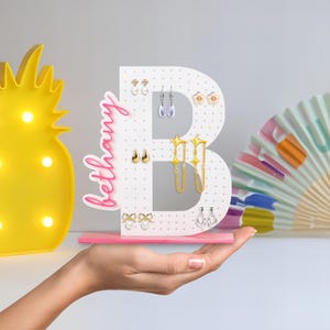 May include: A white, letter "B" shaped earring holder with gold polka dots, holding various earrings. The name "Bethany" is written in pink script on the side. The holder sits on a pink base. A yellow pineapple lamp is in the background.