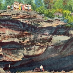 May include: Vintage postcard featuring the Adirondack Natural Stone Bridge near Schroon Lake, N.Y. The image shows a large rock formation with people standing on top. The colors are muted, with shades of brown, green, and blue.