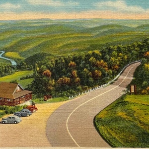 May include: A vintage postcard depicting a scenic view of the Deerfield River Valley from the Mohawk Trail in Massachusetts. The postcard features a winding road leading to a lookout point with a small building and a few cars parked nearby. The valley is lush and green, with a river winding through it. The text "The Deerfield River Valley from Mohawk Trail, Mass. 142" is printed at the top of the postcard.