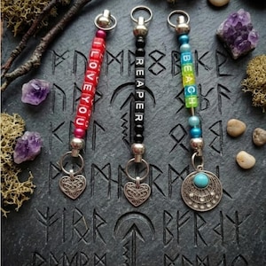 May include: Three keyrings with different designs. One keyring has red beads spelling "I LOVE YOU", another has black beads spelling "REAPER", and the third has beads spelling "BEACH". Each keyring has a heart or circular charm.
