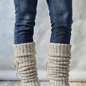 May include: A pair of beige, speckled, chunky knit leg warmers featuring a ribbed texture and a decorative side tie detail. These leg warmers are worn over dark blue denim jeans and black leather ankle boots with a block heel.