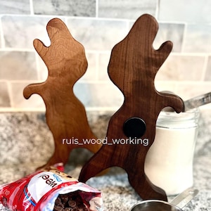 May include: Two wooden reindeer-shaped stands in light and dark brown. A bag of chocolate chips is open, spilling onto the surface. A small metal measuring cup holds more chips. A jar of white powder is in the background.