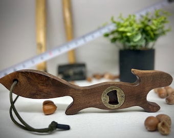 May include: A wooden bottle opener shaped like a squirrel, featuring a brass opener. The opener has a dark brown finish and a green cord attached. Acorns are scattered around the opener.