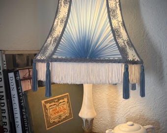 Puede incluir: Una lámpara de mesa de estilo vintage con una pantalla plisada azul y blanca, adornada con bordados florales grises y borlas azules. La lámpara tiene una base de mármol blanco y un soporte de metal color latón. También se ven una tetera y una taza blancas.