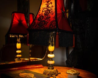 May include: A vintage-style table lamp with a red lampshade adorned with black lace and fringe. The lamp has a decorative base and a cream-colored body. A book and a small brush sit on the wooden table.