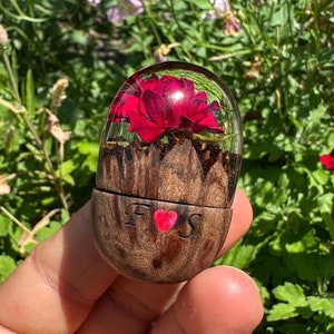 May include: A wooden ring box with a red rose encased in resin. The box is engraved with the letters "F" and "S" with a heart in between.