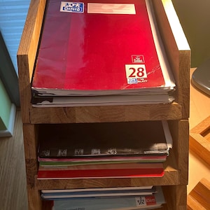 以下が含まれることがあります： 木製の3段のデスクオーガナイザー。ノートや書類が収納されています。一番上の棚には、Oxfordのロゴと数字28が入った赤いノートがあります。他の棚には、様々なノートや書類が収納されています。
