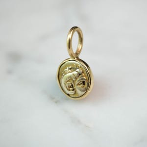 Könnte beinhalten: Ein goldfarbener Anhänger mit einem detaillierten Relief eines Gesichts, möglicherweise einer klassischen Figur. Der Anhänger hat ein kreisförmiges Design mit einer Schlaufe oben für eine Kette. Das Gesicht ist im Profil, mit komplizierten Details. Der Anhänger ist wahrscheinlich aus Gold.