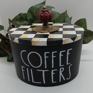 May include: A black, round container with a lid featuring a black and white checkered pattern. The lid is topped with a maroon finial. The words "COFFEE FILTERS" are written in white on the front of the container.