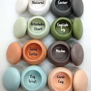 May include: A collection of eight round, smooth containers with matching lids, arranged in four rows and two columns. The colors include white, dark gray, light green, olive green, orange, brown, light blue, and peach, each labeled with text.