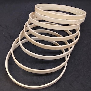 May include: A stack of six circular embroidery hoops made of light-colored bamboo wood. Each hoop features a smooth, unfinished surface and a thin, flexible frame design. The hoops are arranged in a cascading, overlapping formation.