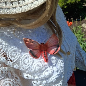 May include: A detailed view of a decorative butterfly pin with a reddish-orange body and wings. The wings have black markings and the pin is attached to a white lace garment. The butterfly is a striking accessory.