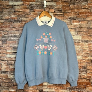 May include: A light blue sweatshirt with a white collar and a floral design. The design includes a basket of flowers, hearts, and two doves. The text "Life's a Good Friend" is also visible.