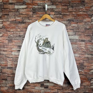 May include: White long-sleeve sweatshirt featuring a detailed graphic of a cougar on a rocky hillside with trees and bare branches. The sweatshirt is hanging on a wooden hanger against a brick wall.