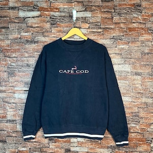 Puede incluir: Sudadera azul marino con la inscripción "CAPE COD MASSACHUSETTS" en blanco y rojo. Los puños y el dobladillo tienen una franja blanca. La prenda está colgada de una percha amarilla.