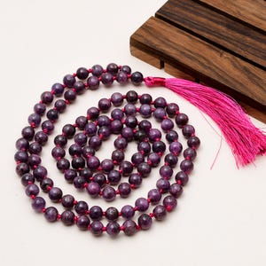 May include: A purple beaded necklace with a bright pink tassel. The beads are a deep purple colour and appear to be made of stone. The necklace is arranged in a spiral pattern on a white surface.