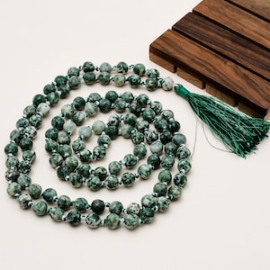 May include: A necklace of green and white speckled beads, possibly gemstones, arranged in a spiral. The beads are strung on a white cord and finished with a green tassel. A small wooden block is in the background.