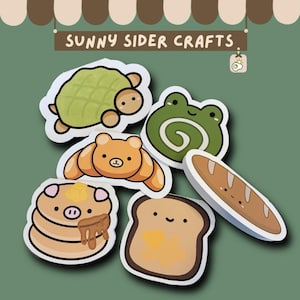 Op de afbeelding: Een verzameling cartoonstickers met een schildpad, kikker, croissant, pannenkoeken, toast en een brood. De stickers hebben een witte rand en een schattig ontwerp. De tekst "SUNNY SIDER CRAFTS" staat bovenaan.