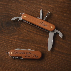 Op de afbeelding: Twee houten Zwitserse zakmessen met diverse gereedschappen, waaronder een flesopener, mes en kurkentrekker. Eén mes is gegraveerd met de naam "Leon", en de andere met de tekst "Tu seras riche... d'un Pere".