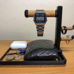 May include: A black and cork watch stand with a digital watch, a black leather wallet, a white AirPod case, keys, and two coins. The stand has two hooks for rings.