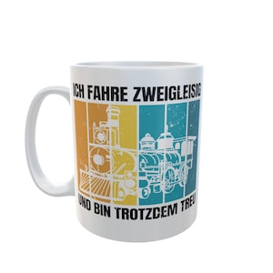 Könnte beinhalten: Weiße Keramiktasse mit farbenfrohem Zugdesign. Das Design zeigt eine Vintage-Zugillustration in Orange, Gelb und Blau mit deutschem Text. Die Tasse hat einen gebogenen Griff und ein glänzendes Finish.