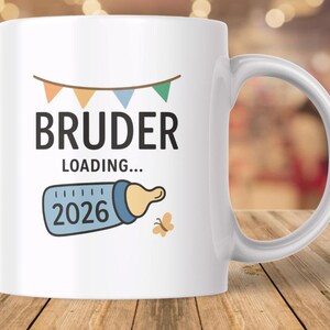 Może przedstawiać: Biały ceramiczny kubek z napisem "BRUDER LOADING..." i grafiką butelki dla dziecka z rokiem "2026". Kubek posiada również baner z pomarańczowymi, żółtymi i zielonymi flagami oraz małego motyla.